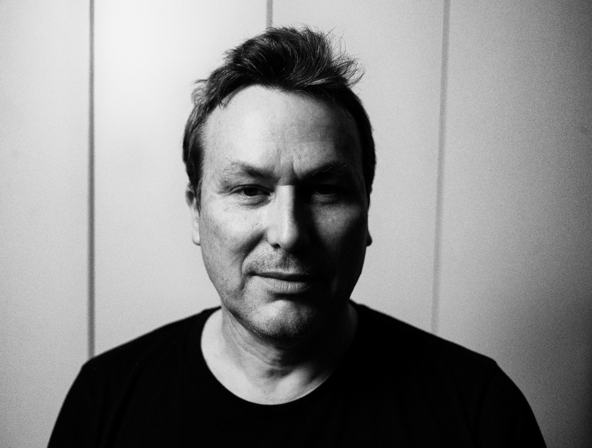 A black and white photo of a man with a subtle moutache smiling at the camera while wearing a black shirt in front of a wall.