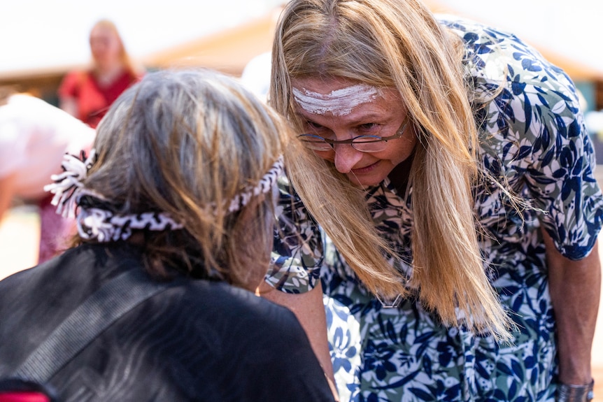 A woman with blonde hair and glasses leans down to talk to an Indigenous person.