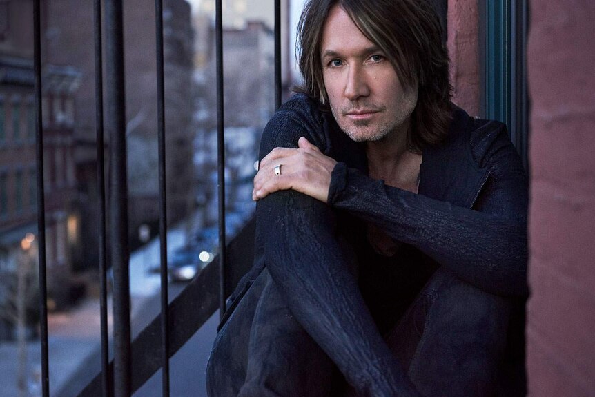 Promotional photo of singer and songwriter Keith Urban sitting with crossed legs and arms, looking at the camera.