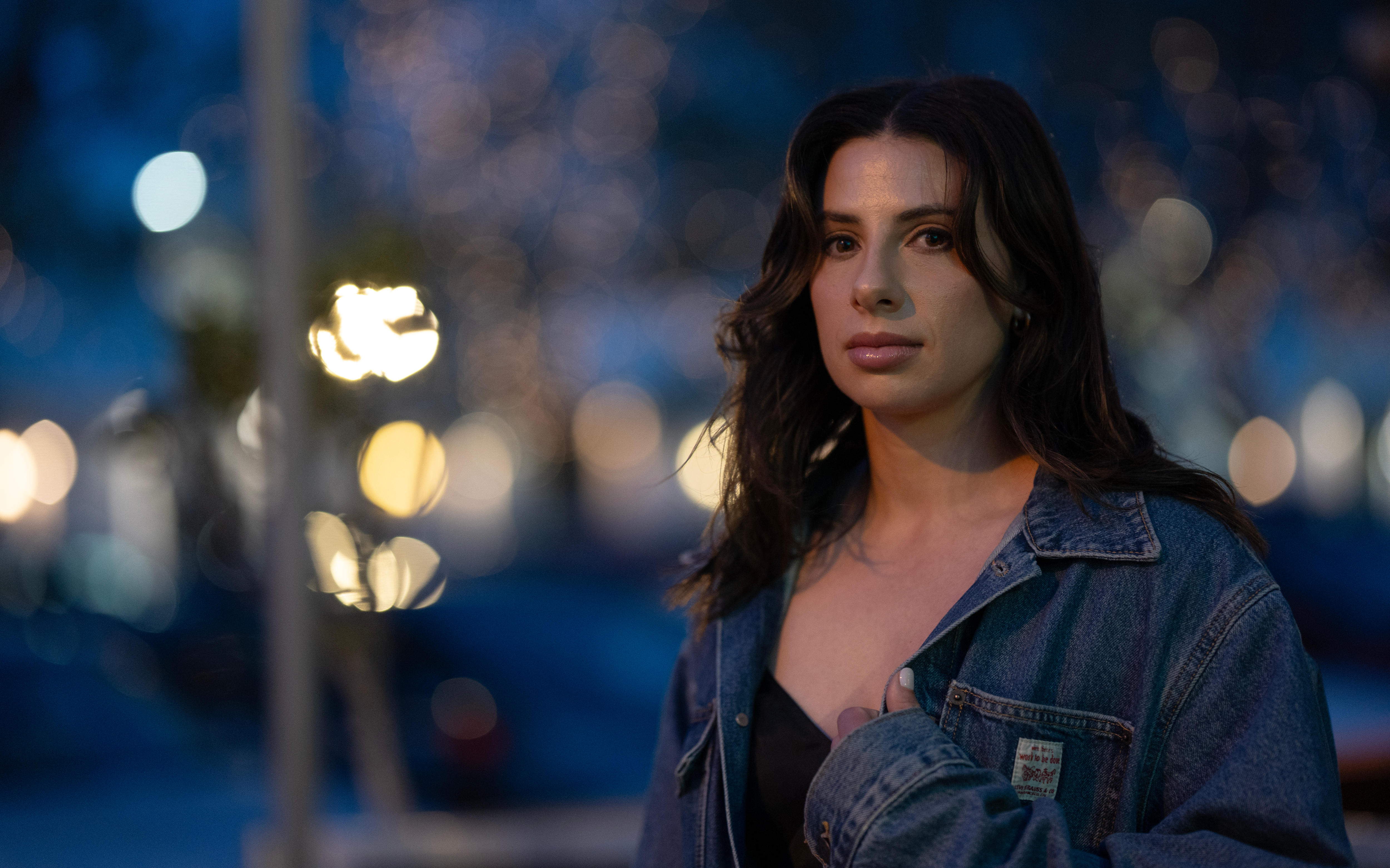 Woman wearing black top and denim jacket with long brown hair standing on a street with city lights behind her.