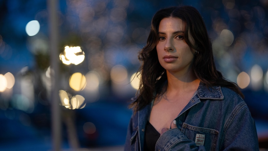 Woman wearing black top and denim jacket with long brown hair standing on a street with city lights behind her.