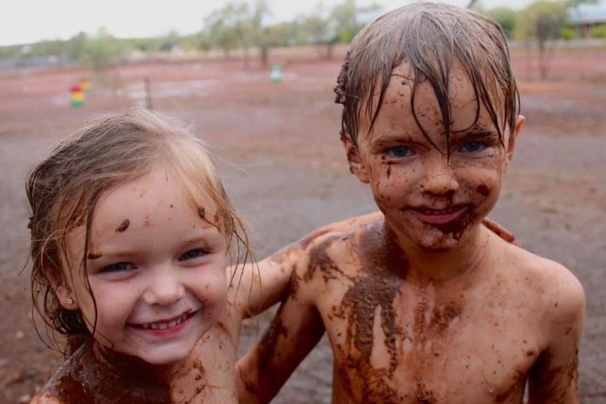 A boy and a girl stand covered in mud with their arms around eachother