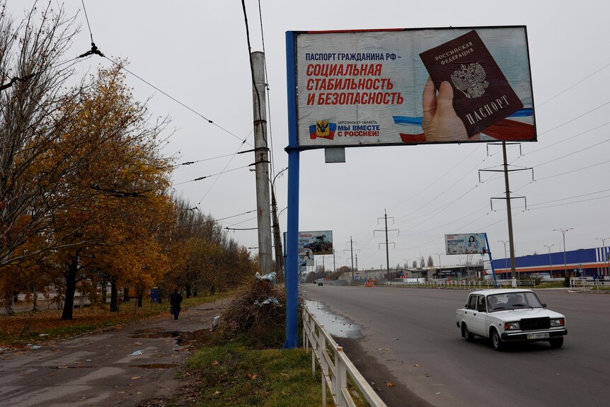 A billboard with Russian script and a picture of a passport hangs over a freeway in a bleak-looking town.