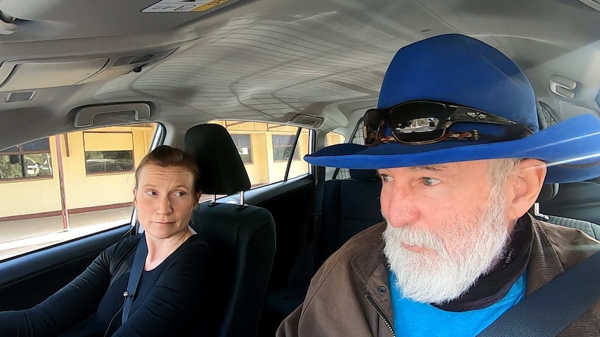 blond haired woman driving car speaking to man wearing a bright blue cowboy styled hat in front passenger seat