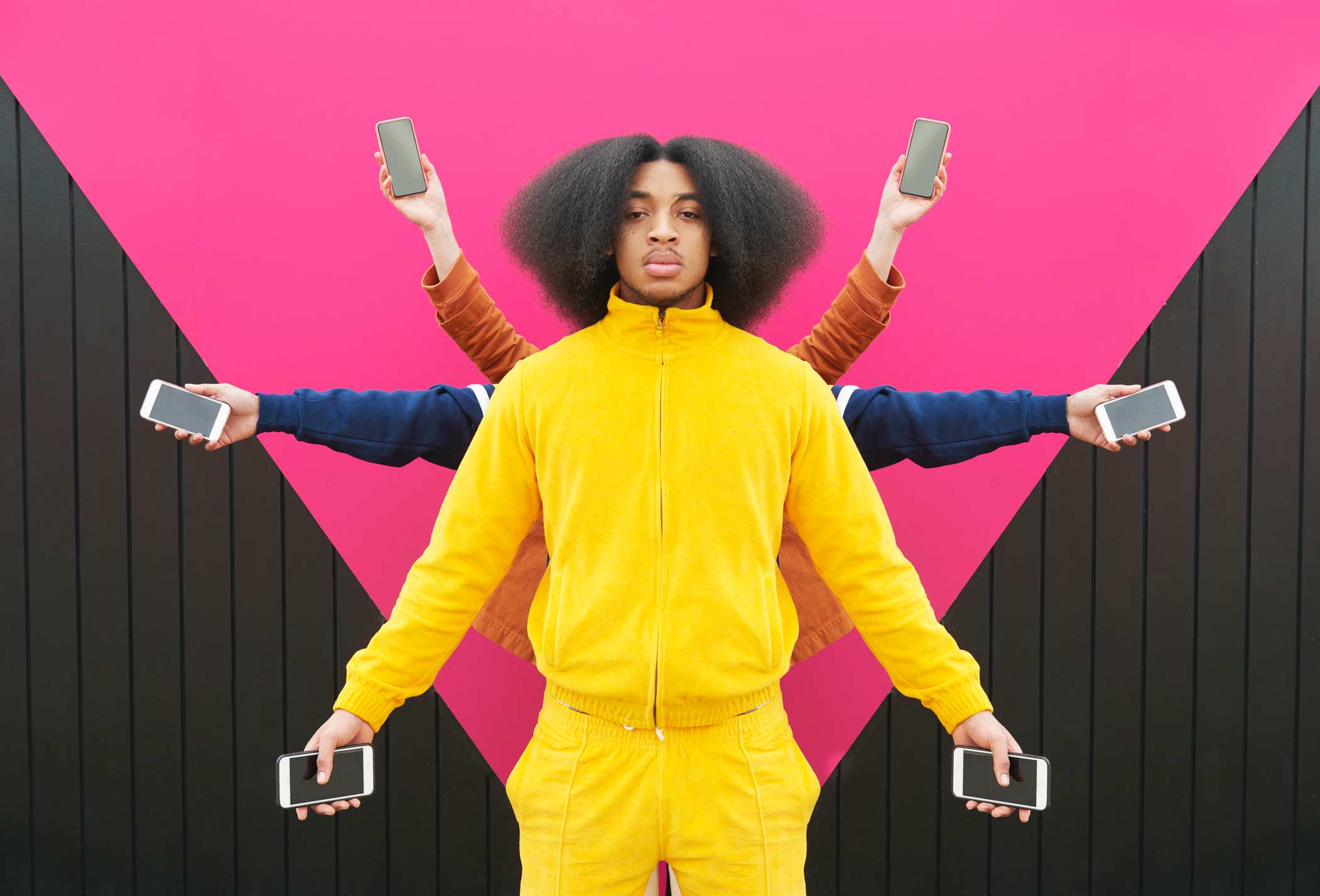 Young man in bright yellow jumpsuit holding phones in both hands. Behind him arms are holding phones creating a star-like shape.