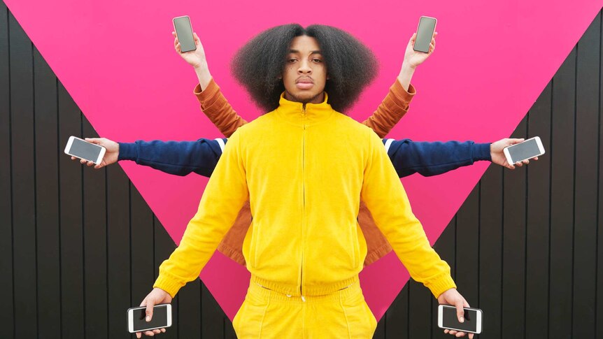 Young man in bright yellow jumpsuit holding phones in both hands. Behind him arms are holding phones creating a star-like shape.