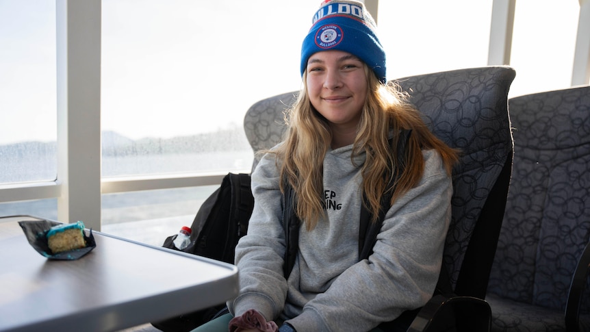 A young woman with long blonde hair and a Western Bulldogs beanie sits at a table on a ferry.