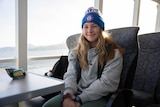 A young woman with long blonde hair and a Western Bulldogs beanie sits at a table on a ferry.