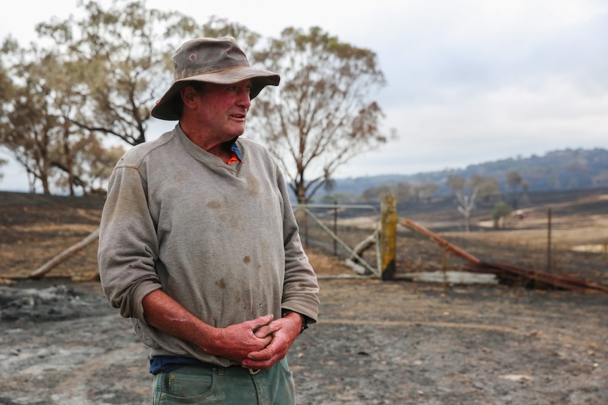 A man in a dirty grey jumper and dirty hate standing on and next to burnt out paddocks.