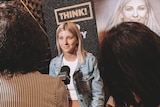 A woman stands being interviewed by two people in front of a number of posters. 