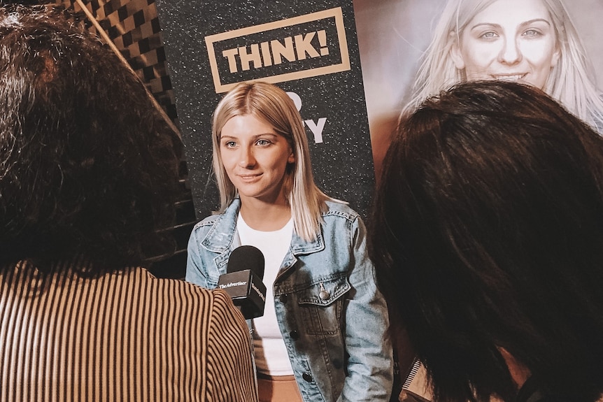 A woman stands being interviewed by two people in front of a number of posters. 