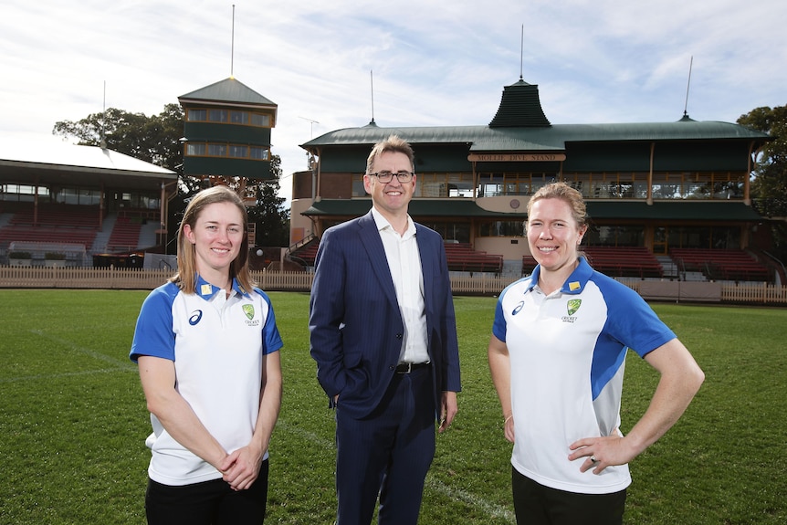 A man in a blue suit and open-neck white shirt stands between two female cricketers dressing in training gear