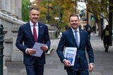 Two men wearing navy suits holding budget papers walk outside on a city street