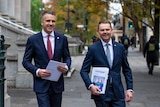 Two men wearing navy suits holding budget papers walk outside on a city street