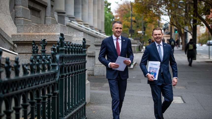 Two men wearing navy suits holding budget papers walk outside on a city street