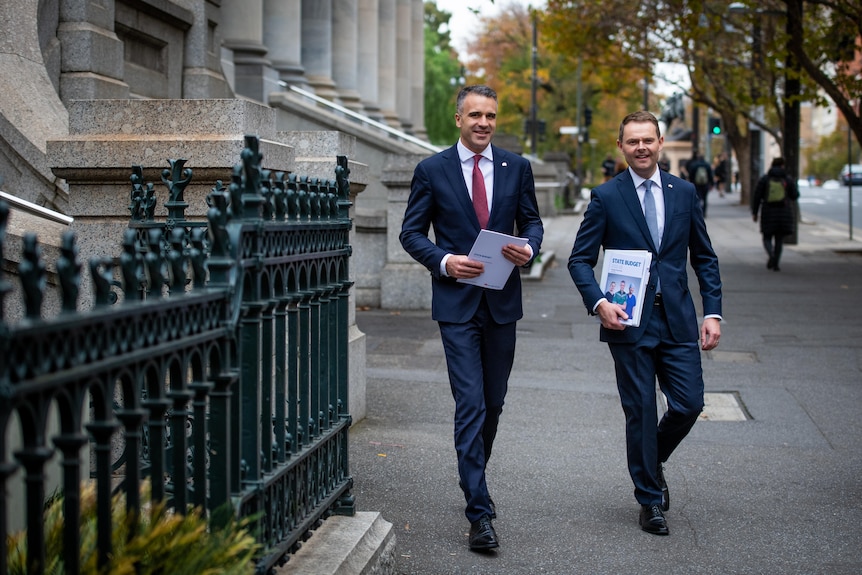 Two men wearing navy suits holding budget papers walk outside on a city street