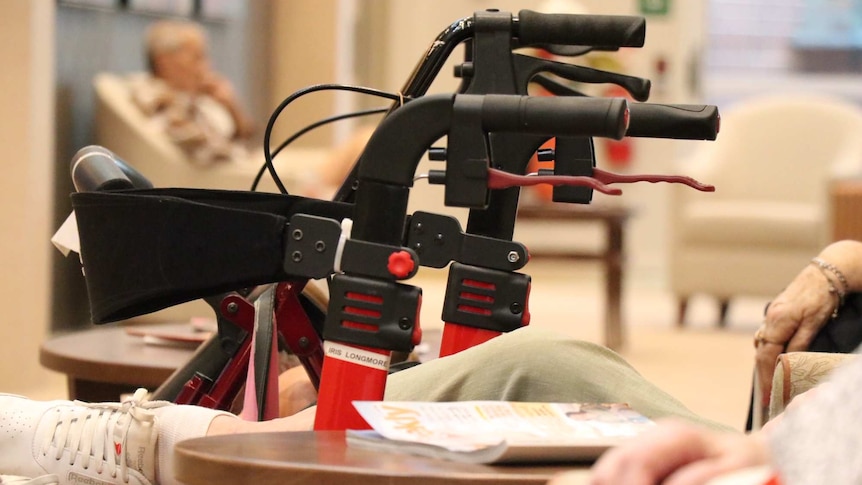 A Zimmer frame in focus with a group of aged care residents (unidentifiable) sitting around it.