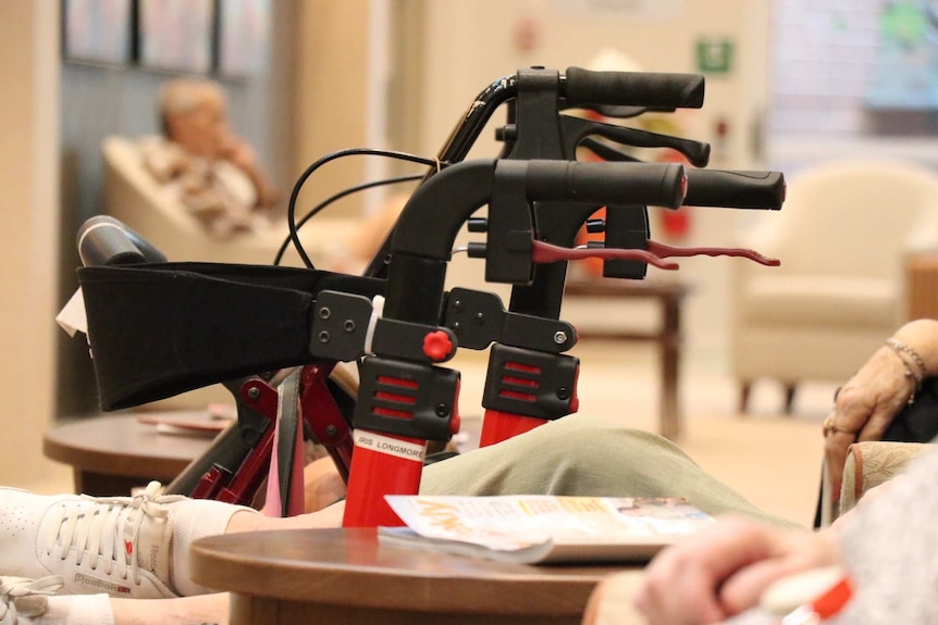 A Zimmer frame in focus with a group of aged care residents (unidentifiable) sitting around it.