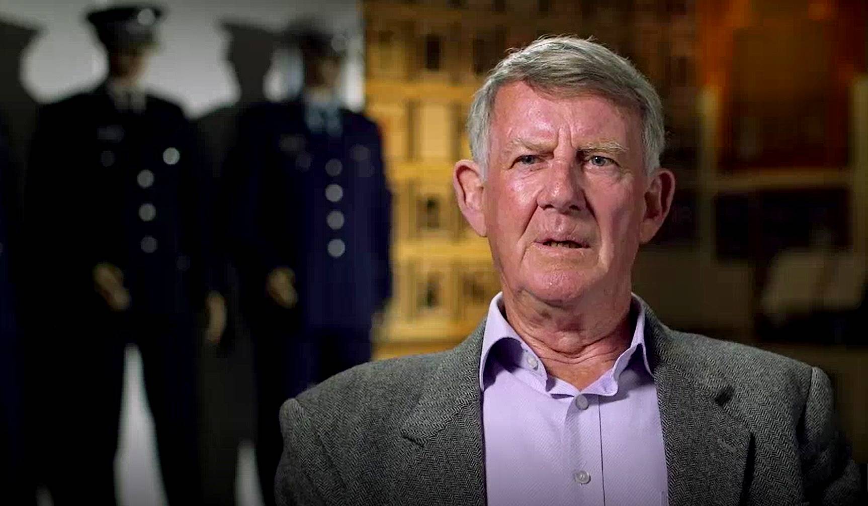 A head shot of an older grey-haired man, wearing a purple shirt, gray jacket.  Mannequins of police behind him.