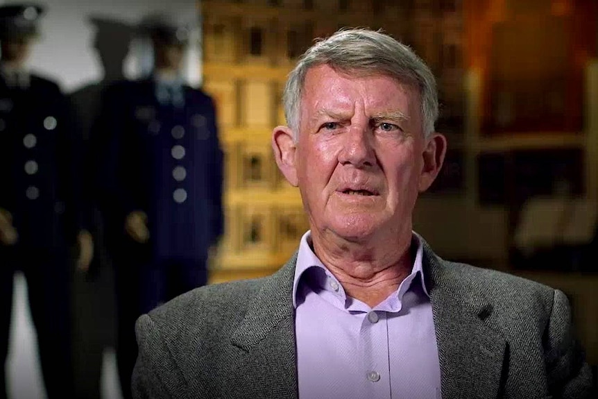 A head shot of an older grey-haired man, wearing a purple shirt, gray jacket.  Mannequins of police behind him.