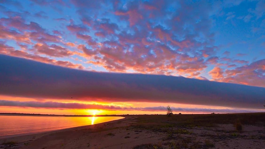 A sunset over a beach, with blue sky, pink clouds, and the sun rising on the left.