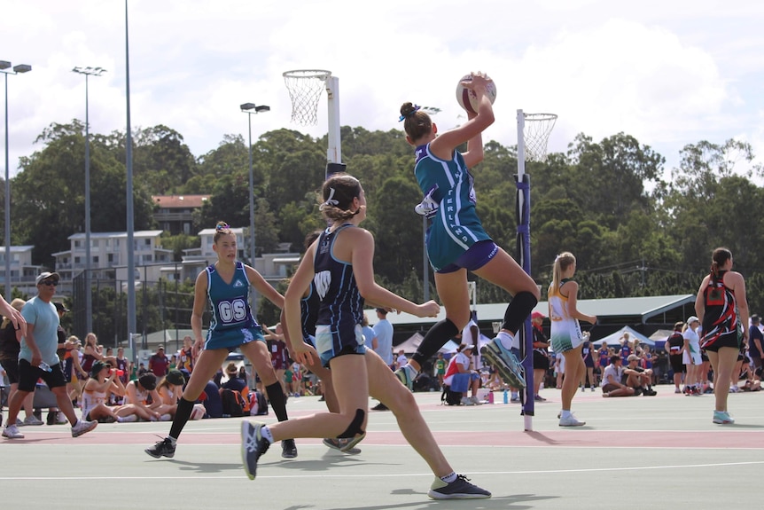 A female netball player in blue leaps with ball in hand with other players run beside in front of onlookers.