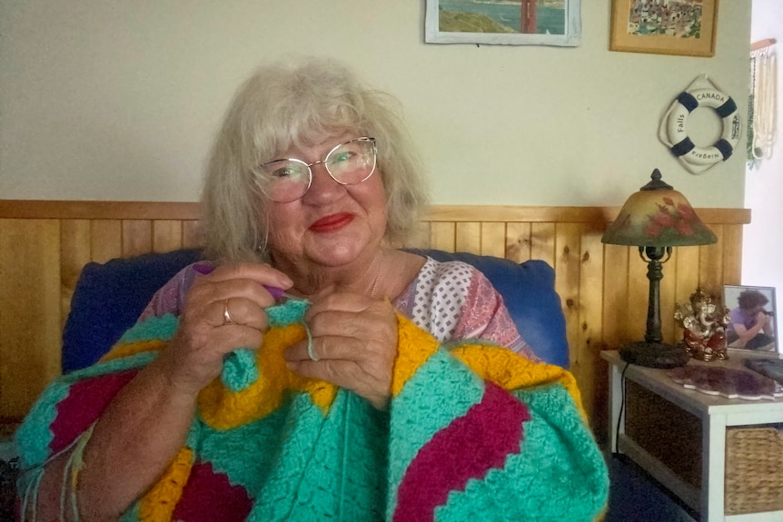 A woman with grey hair, glasses, red lipstick holds crochet hook and crochet rug in red, green, yellow