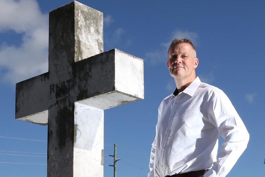 Close up of a man looking serious standing beside a cross in a graveyard.