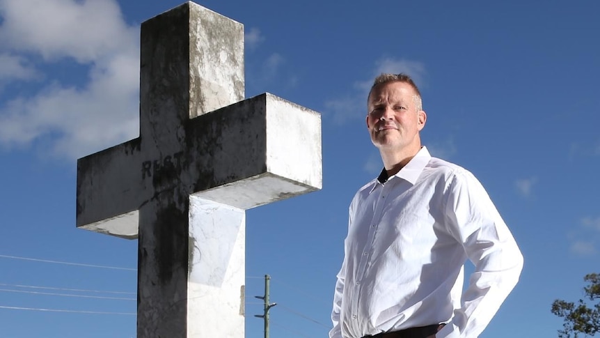 Close up of a man looking serious standing beside a cross in a graveyard.