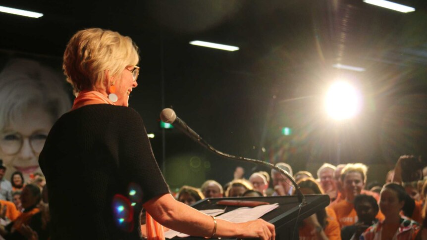 Helen Haines stands at a podium under a bright light in a room filled with supporters dressed in orange.