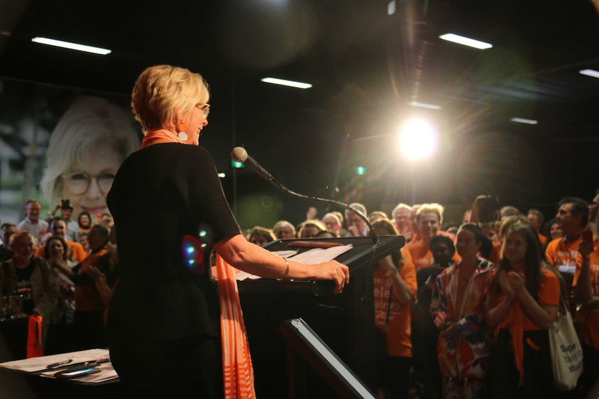 Helen Haines stands at a podium under a bright light in a room filled with supporters dressed in orange.