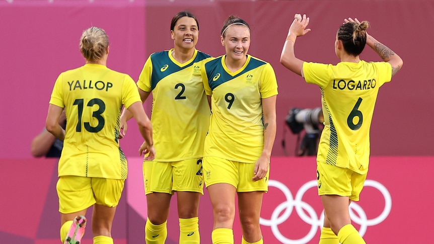 Sam Kerr and Caitlin Foord smile while teammates run up to high five them