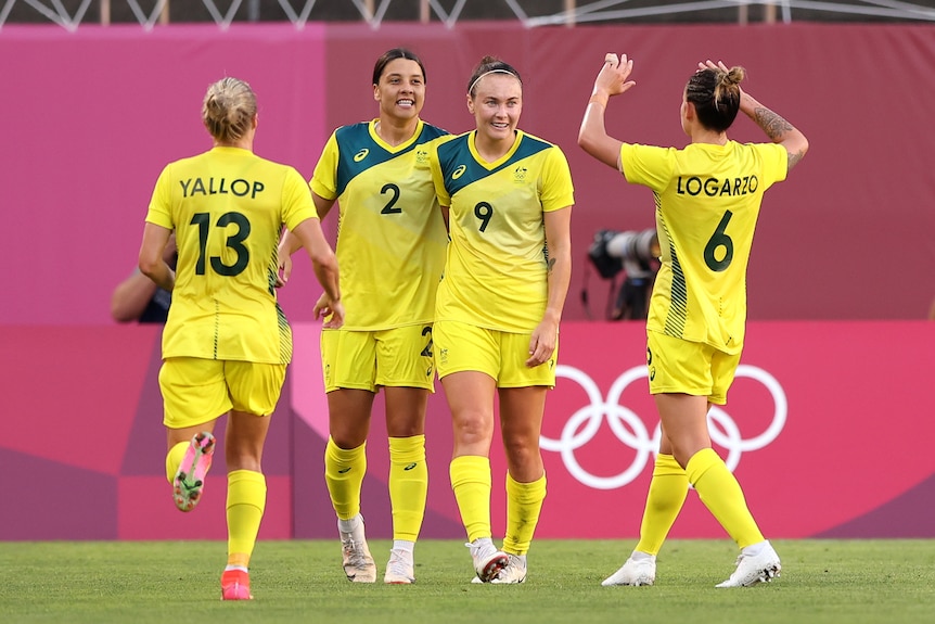 Sam Kerr and Caitlin Foord smile while teammates run up to high five them