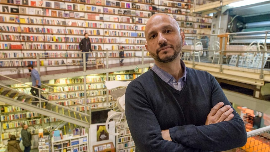 Man with bald head and beard with goatee standing inside bookstore with walls lined in shelving with books.