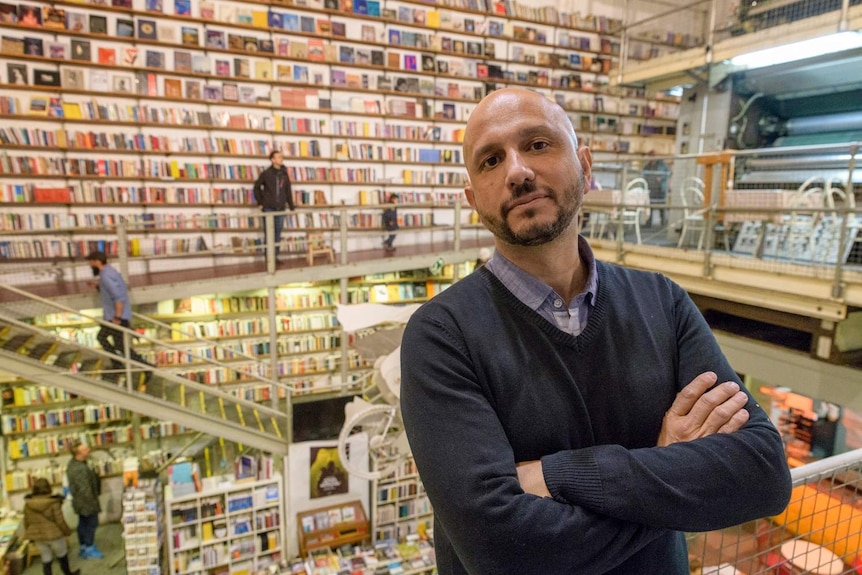 Man with bald head and beard with goatee standing inside bookstore with walls lined in shelving with books.