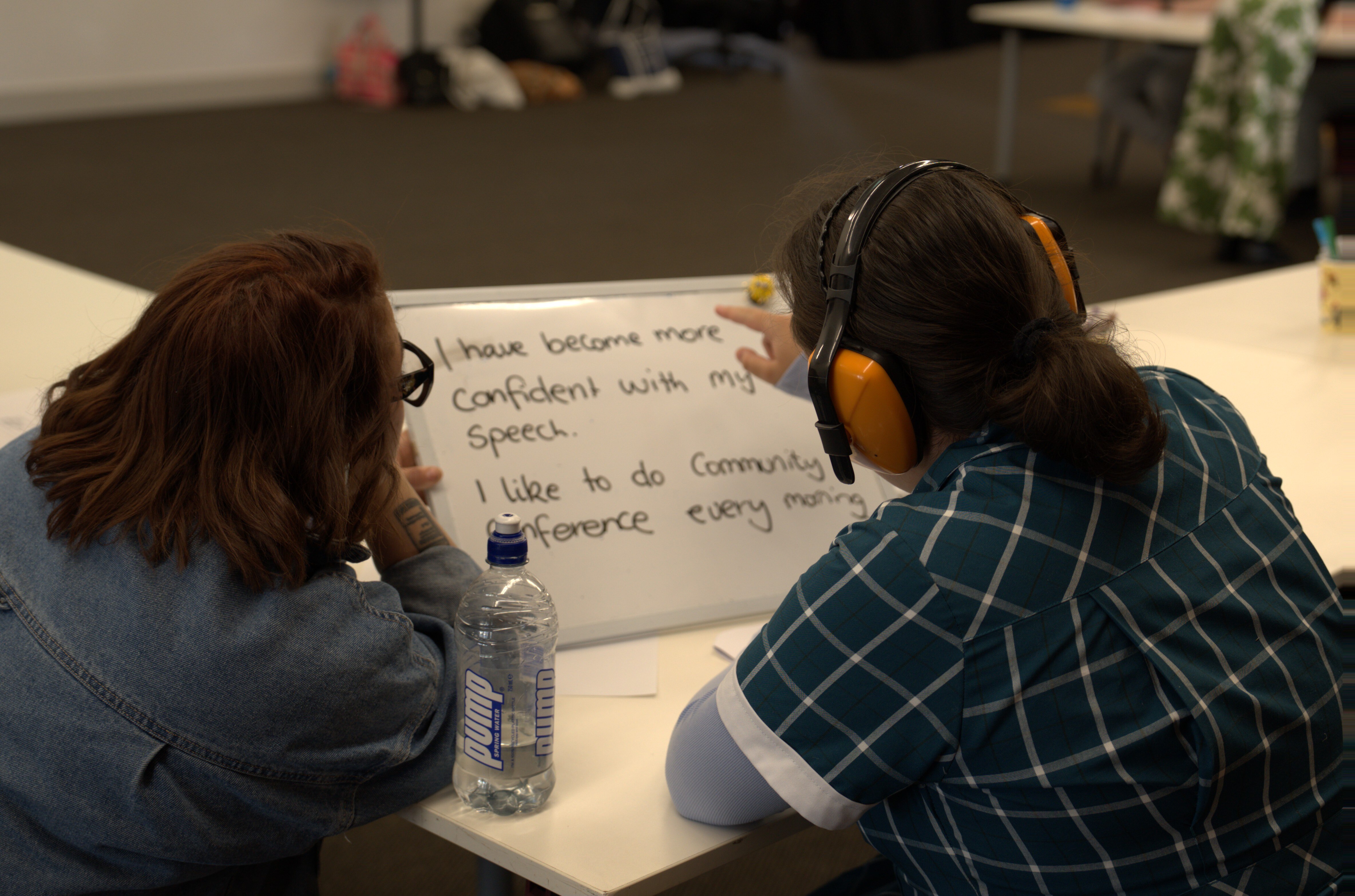 Duas pessoas sentadas em uma mesa segurando um pequeno quadro negro com as palavras "Fiquei mais confiante com meu discurso.".