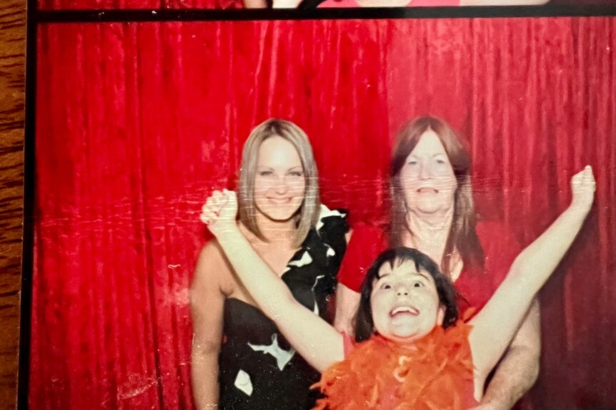 A photo booth picture of a little girl making funny faces with two amused women behind her. 