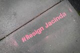 Pink graffiti painted on a cement footpath reads "#Resign Jacinda"