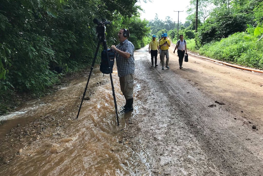 Cameraman and camera on tripod standing in stream of muddy water alongside road.
