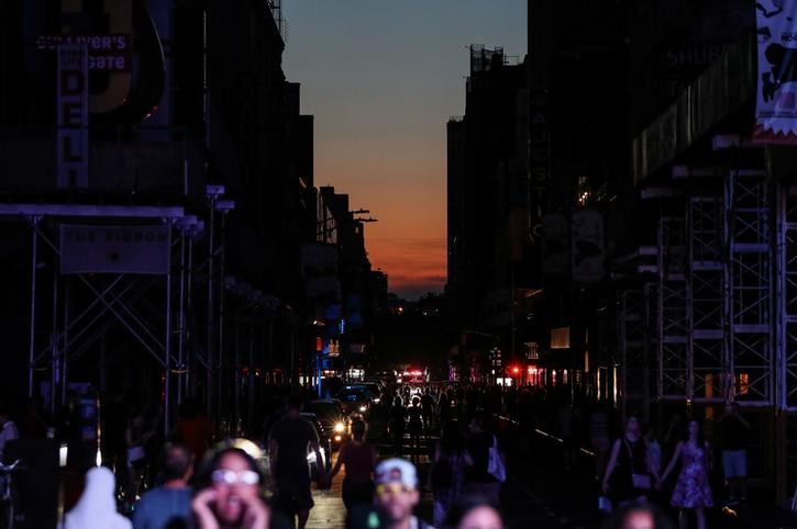 Buildings are plunged in darkness on the left and right with people walking in front of camera and sun setting in distance.