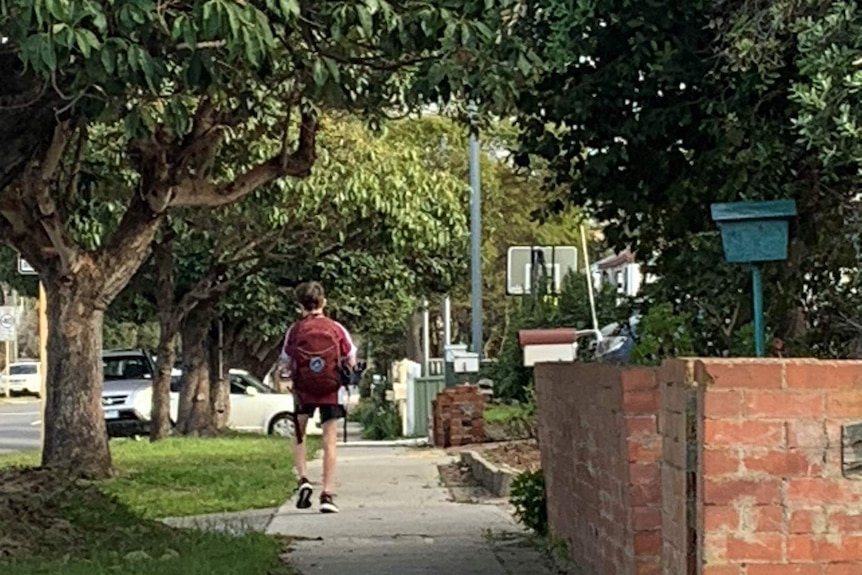 A boy with a maroon school backpack walks down a suburban footpath past houses and trees