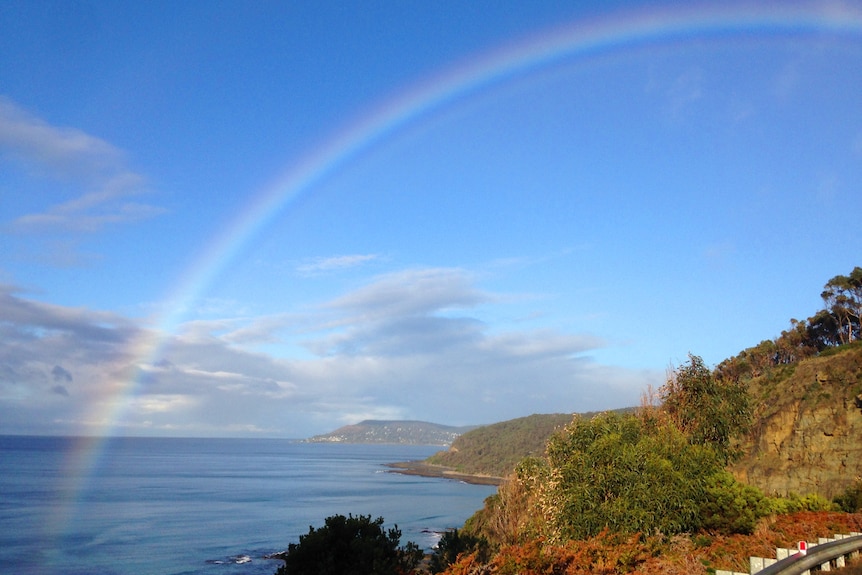 A large rainbow over the ocean on a blue sky day. Two headlands are seen in the distance.