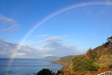 A large rainbow over the ocean on a blue sky day. Two headlands are seen in the distance.