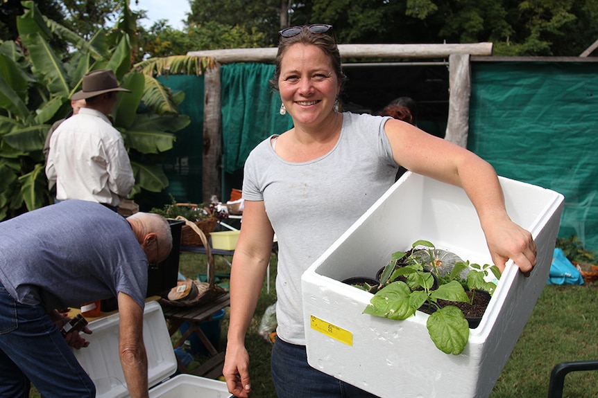 A woman in a grey shirt and jeans holding onto a white foam box carrying a pot plant, a whole pumpkin and other produce.