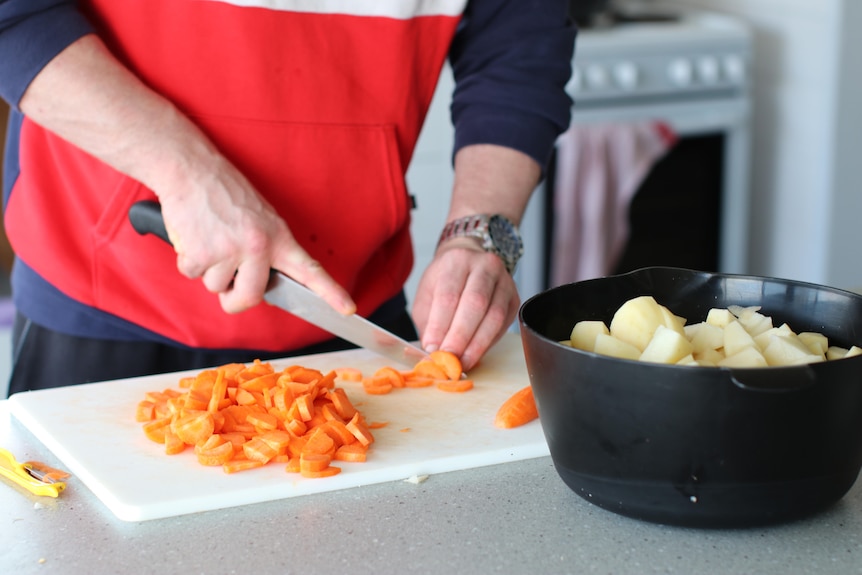 Close up of man's hands cutting carrots on cutting board with pot of potatoes nearby