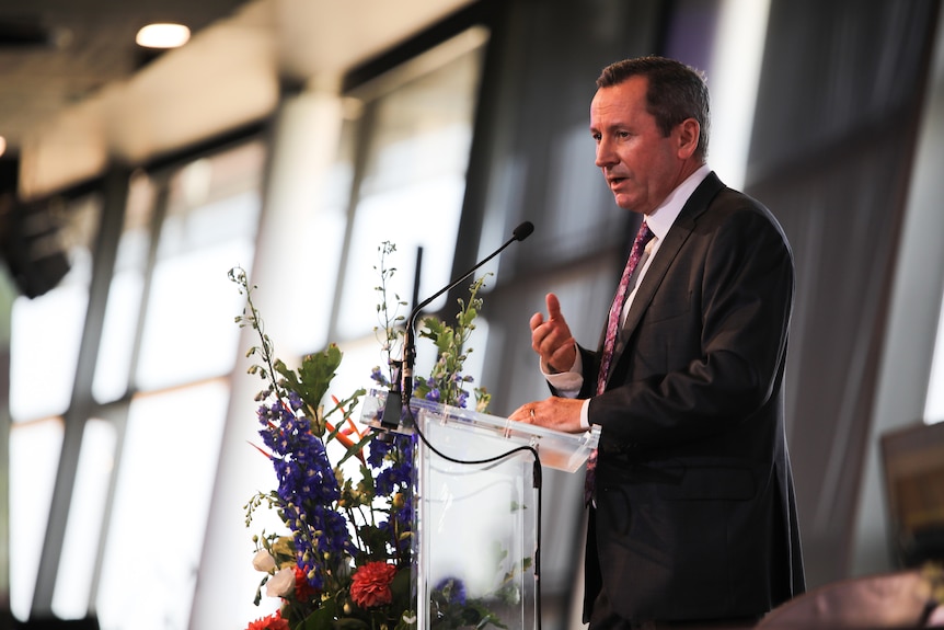 WA premier Mark McGowan speaks at a lectern with some flowers in front of him.