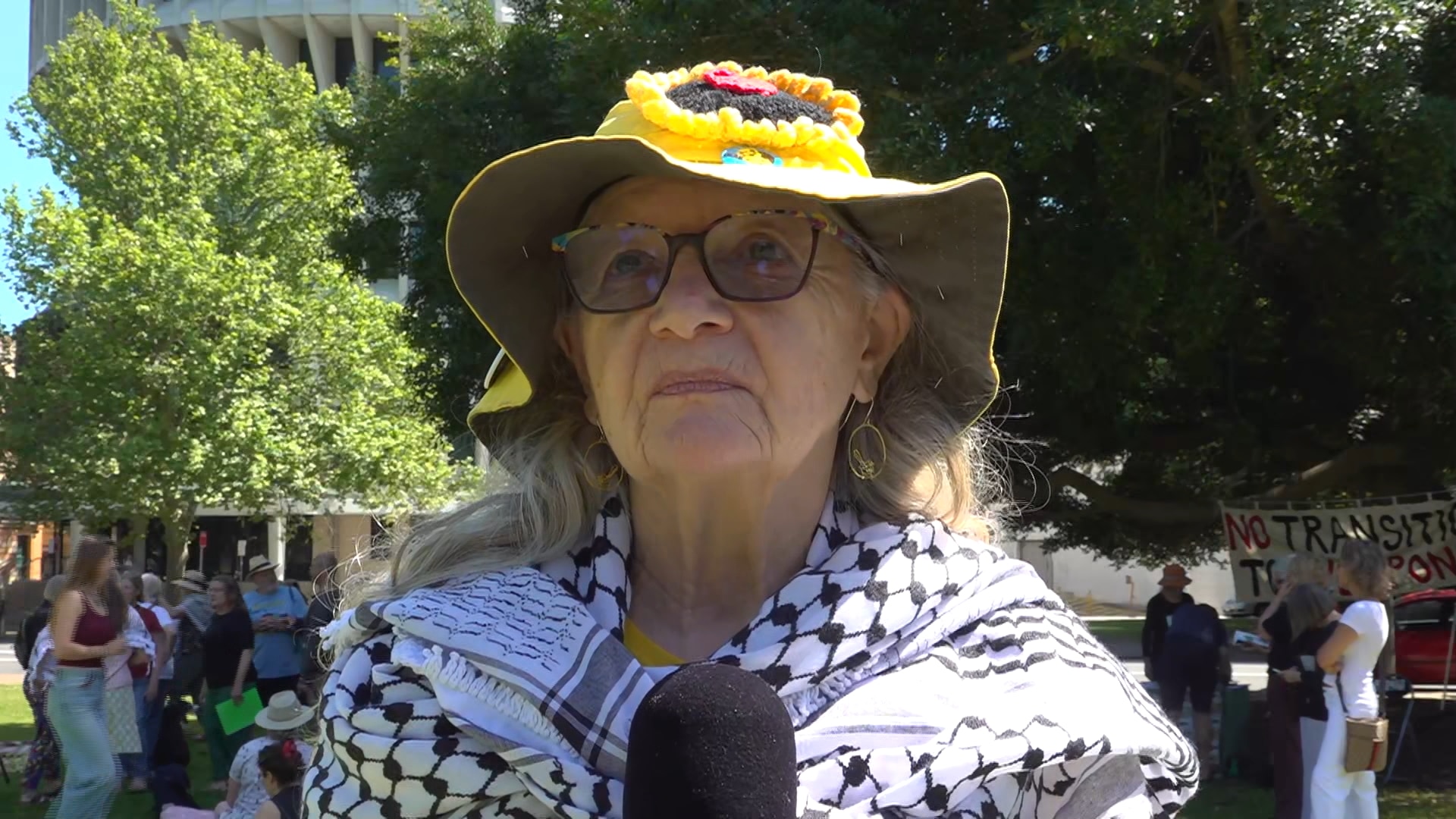 An older woman wearing a scarfe, glasses and a widebrimmed hat outside speaking into a microphone.
