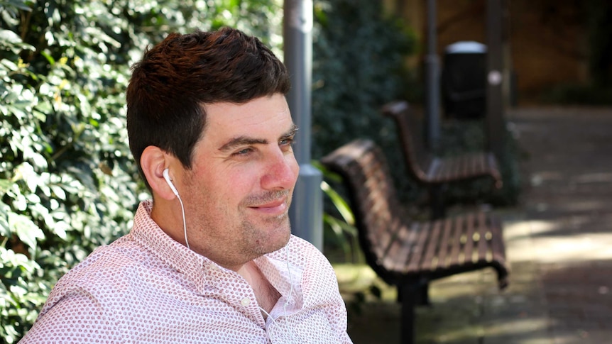 A smiling man sits on a park bench listening through headphones.