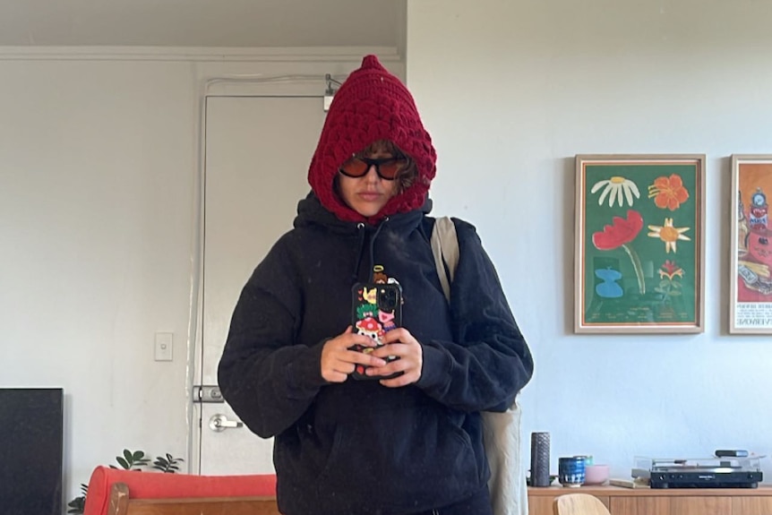 Carmen wears an oversized jacket, a red knit balaclava and baggy pants in a mirror selfie.