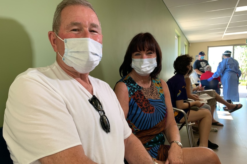 A couple sits in a busy clinic waiting room with face masks on.